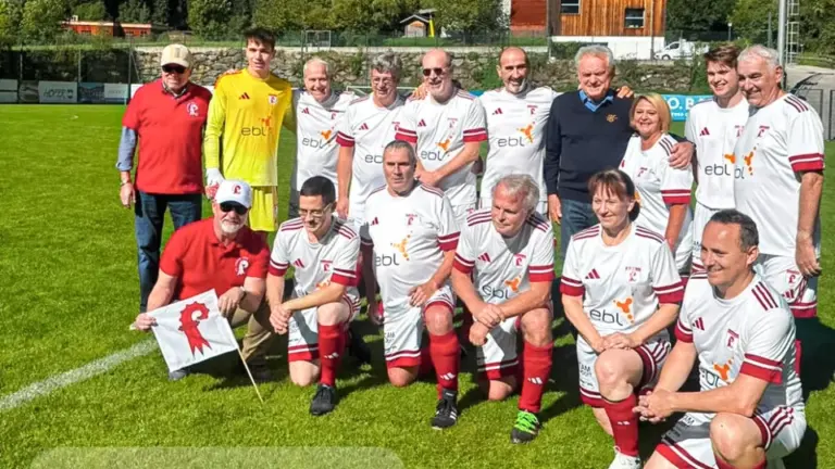 Fussballmannschaft in weissen Trikots posiert gruppenweise auf einem Sportplatz vor Bergkulisse.