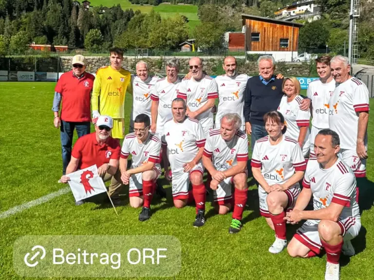 Fussballmannschaft in weissen Trikots posiert gruppenweise auf einem Sportplatz vor Bergkulisse.
