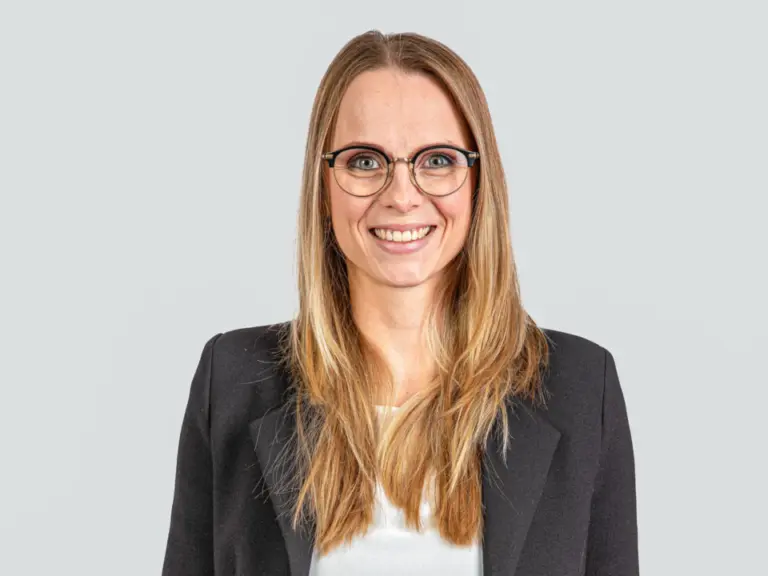 Lächelnde Frau mit Brille und langen blonden Haaren, in schwarzem Blazer und weisser Bluse.