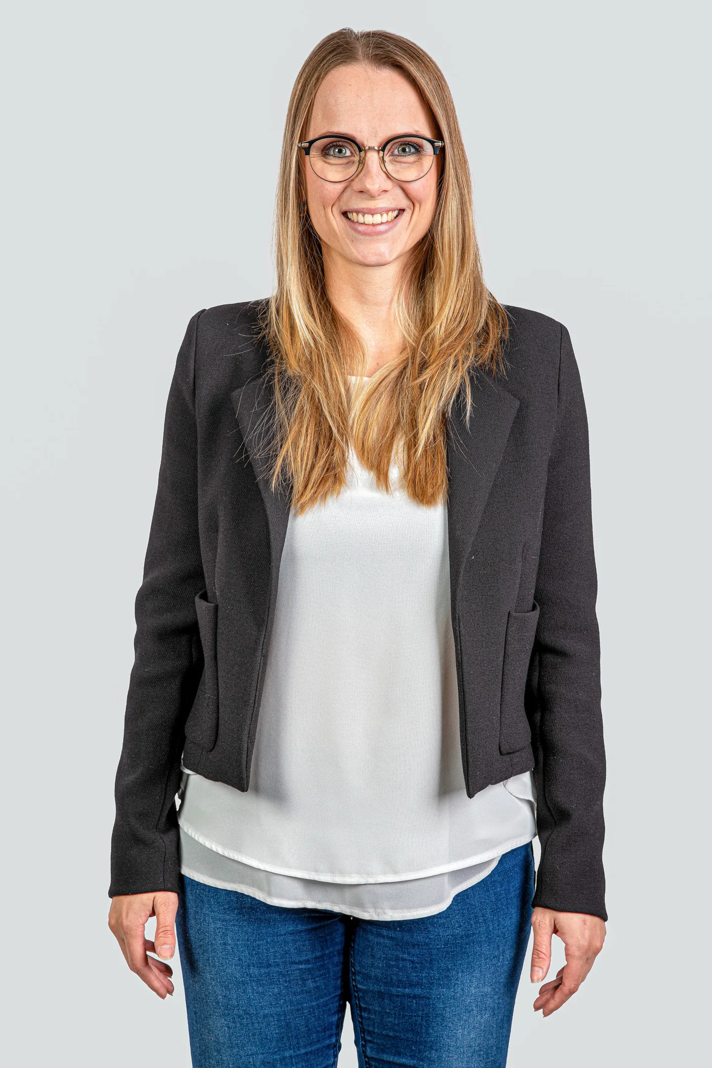 Lächelnde Frau mit Brille und langen blonden Haaren, in schwarzem Blazer und weisser Bluse.