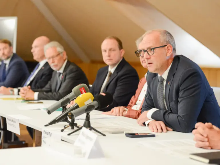 Mehrere Männer in Anzügen sitzen nebeneinander an einem Tisch mit Mikrofonen bei einer Pressekonferenz.
