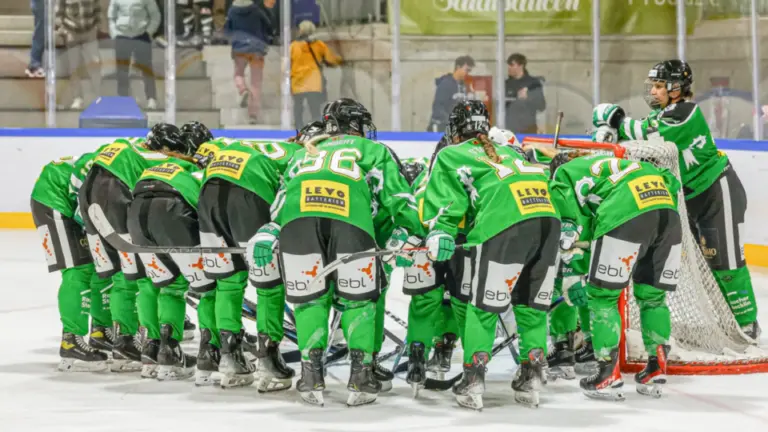 Eishockeyspielerinnen in grünen Trikots bilden vor dem Spiel einen Kreis auf dem Eis.
