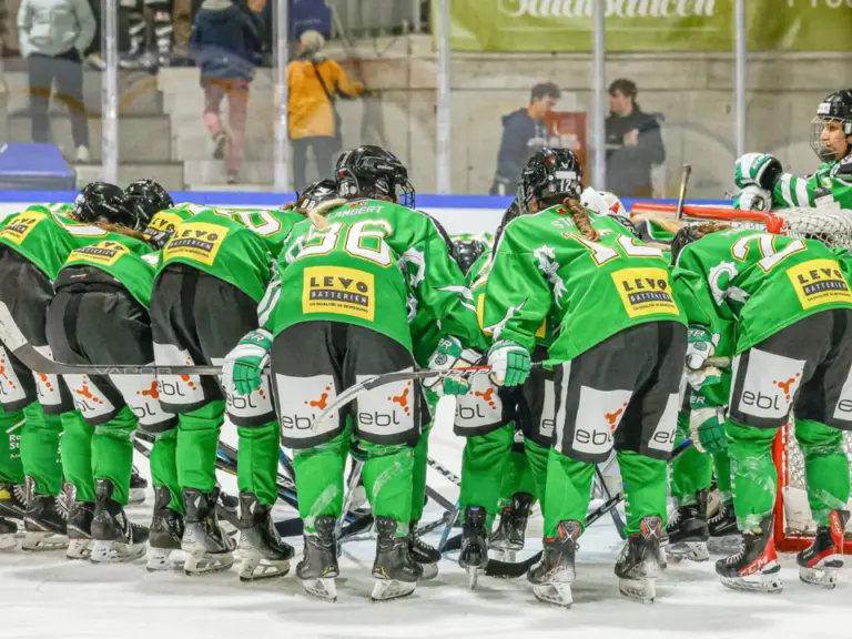 Eishockeyspielerinnen in grünen Trikots bilden vor dem Spiel einen Kreis auf dem Eis.
