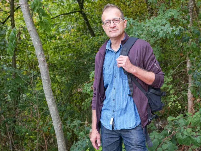 Ein Mann mit Brille und Rucksack steht lächelnd vor dichtem Waldgebüsch.